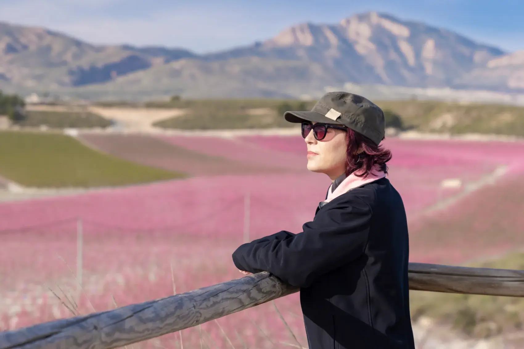 Cicloturistas recorren la Vía Verde de la Floración de Cieza, sobre el antiguo trazado ferroviario, entre campos de frutales de hueso en flor teñidos de rosa, con la silueta de las sierras y cielo de nubes al fondo