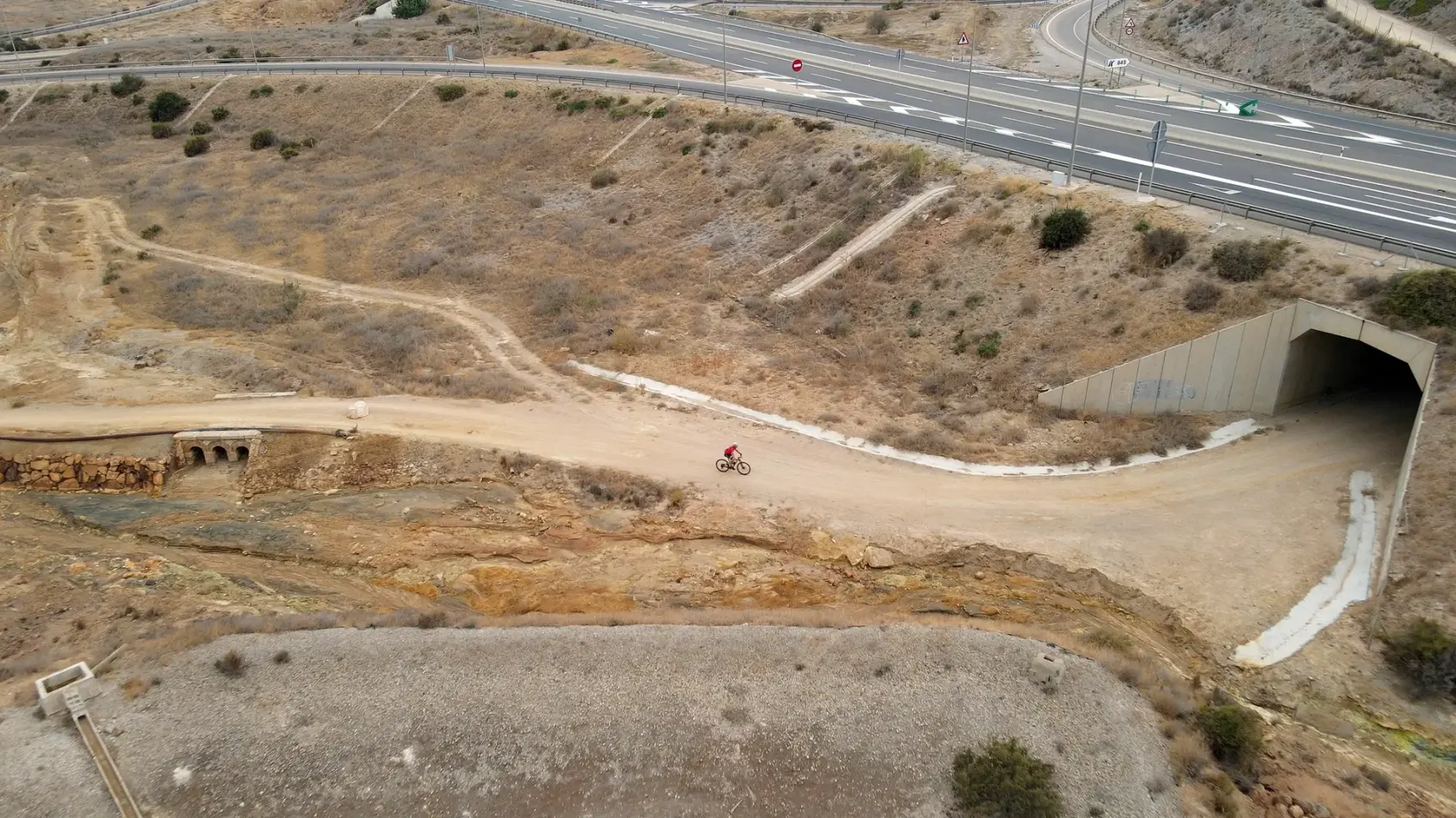 Camino de tierra compactada de la Vía Verde de Mazarrón descendiendo entre los cabezos ocres y rojizos del antiguo Coto Minero de San Cristóbal bajo un cielo azul